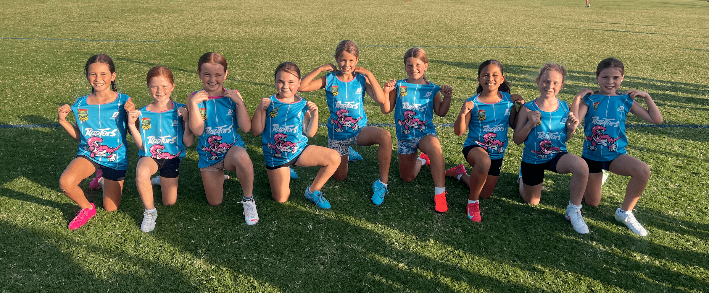 Nine young girls in matching blue Raptors jerseys kneel and smile on a grassy field.